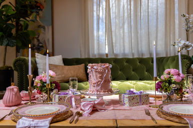 Pink-themed tablescape with ornate pink cake on a glass stand, lit taper candles, floral bouquets and wrapped gifts on a wooden dining table set before a green velvet tufted sofa