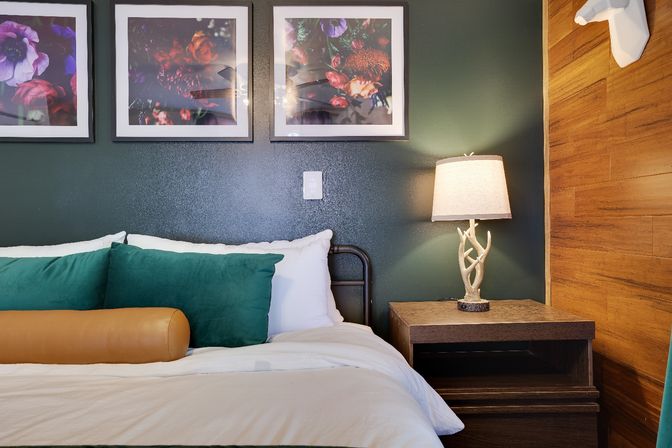 Cozy modern bedroom interior with a deep green accent wall, three framed floral prints, white bedding with green pillows and a tan bolster, wooden nightstand topped by an antler-style lamp, and warm wood-paneled accent wall.