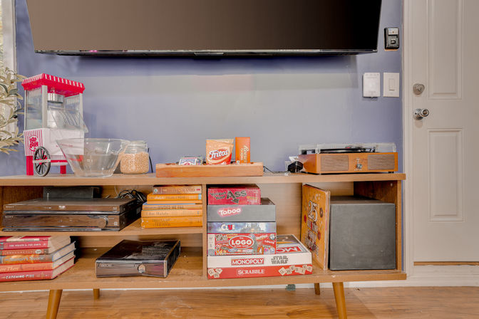 Mid-century wooden media console under a wall-mounted TV in a living room, with a retro red popcorn machine, clear snack bowls and chips, a turntable, stacked board games, books and a black subwoofer — cozy family game-night setup.