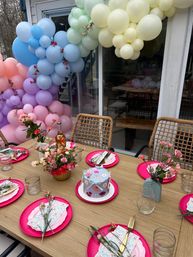 Backyard patio party table with pastel balloon arch, bright pink plates, floral centerpieces, wicker chairs and a decorated mini cake.