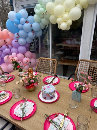 Backyard patio party table with pastel balloon arch, bright pink plates, floral centerpieces, wicker chairs and a decorated mini cake.