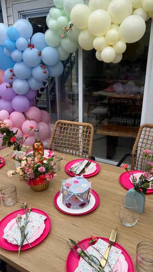 Backyard patio party table with pastel balloon arch, bright pink plates, floral centerpieces, wicker chairs and a decorated mini cake.