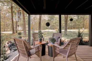 Cozy covered porch with four wicker chairs around two wood‑top tables, potted plants, hanging greenery and a wind chime, overlooking a tree‑lined yard and quiet country road.