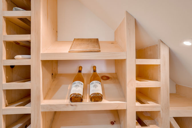 Cozy light-wood wine rack tucked under a sloped ceiling with two wine bottles resting on a shelf and a leather-bound book above, bathed in warm recessed light