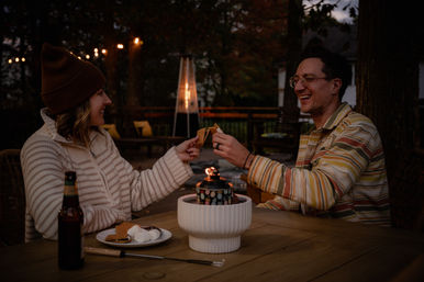 Two friends on a backyard deck at dusk, smiling and toasting s'mores over a tabletop fire pit with string lights and a patio heater in the background.