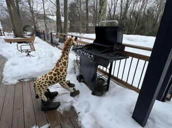 Playful stuffed giraffe wearing boots standing by a gas grill on a snow-covered backyard deck with leafless trees and nearby houses in a winter patio scene