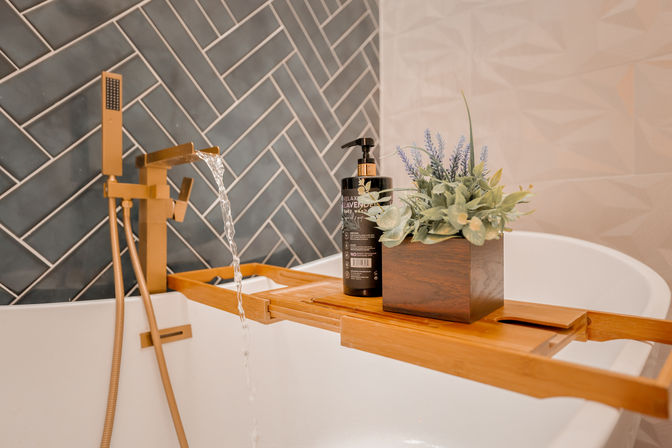 Spa-like modern bathroom: brass waterfall faucet filling a white freestanding tub, wooden bath tray with a black lavender soap dispenser and potted lavender in front of dark gray herringbone tile.