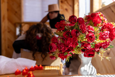 Vibrant red rose bouquet in a white vase on a wooden spa table with tea lights, blurred massage treatment and cozy cabin-style interior in the background