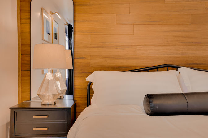 Cozy modern bedroom with warm wood accent wall, crisp white bedding on a black metal bed frame, black bolster pillow, dark nightstand topped with a faceted table lamp and tall arched mirror