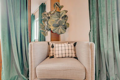Cozy beige armchair with plaid tassel pillow framed by green velvet curtains and a fiddle-leaf fig plant in a modern living room nook