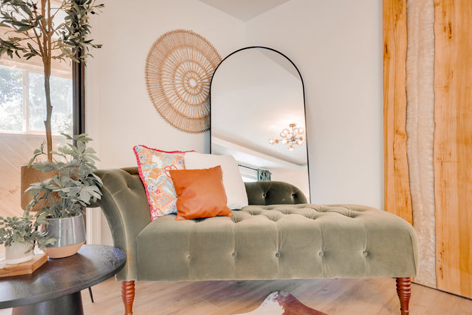 Cozy boho-chic living room corner with an olive-green tufted chaise lounge, colorful patterned and leather pillows, arched black-framed floor mirror, round woven wall decor, potted plants, wooden side table and warm natural light.