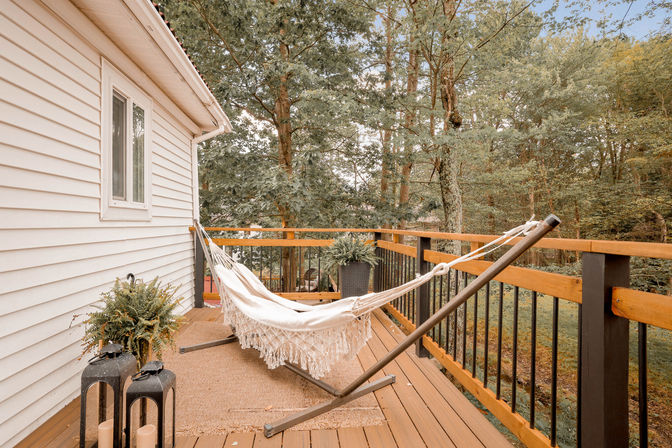 Cozy cream hammock with fringe on a wooden backyard deck beside a white house, overlooking a leafy forest with potted ferns and decorative lanterns.