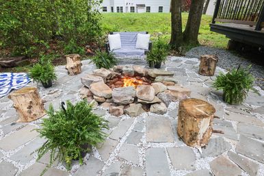 Cozy backyard flagstone patio with a burning stone fire pit, rustic log stools, potted ferns and a striped loveseat with pillows.