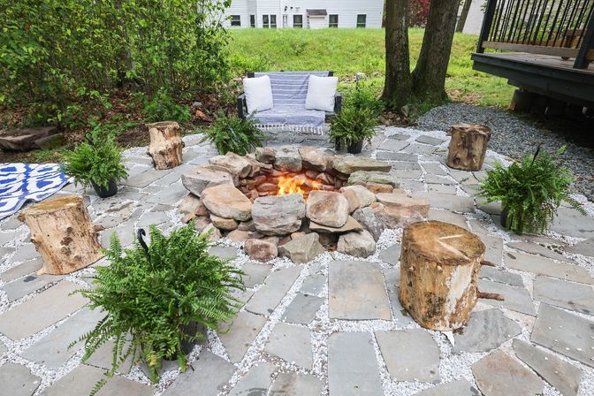 Cozy backyard flagstone patio with a burning stone fire pit, rustic log stools, potted ferns and a striped loveseat with pillows.