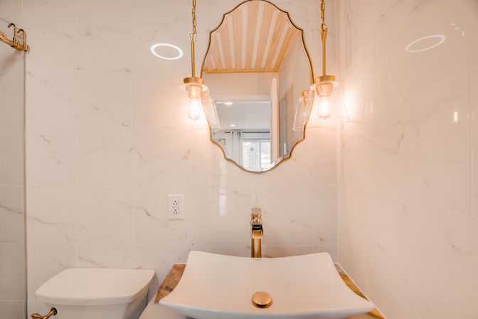 Modern white marble-tiled bathroom vanity with a square vessel sink, gold faucet, scalloped mirror and two glowing glass pendant lights
