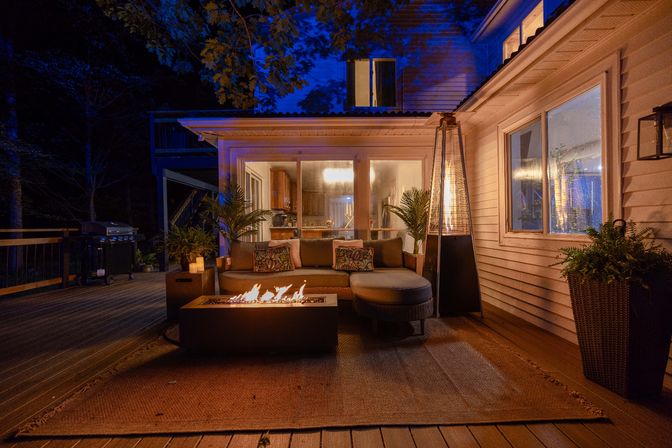 Cozy nighttime backyard deck and outdoor living space with a glowing modern fire pit, cushioned sectional sofa on a rug, tall patio heater and potted plants beside sliding glass doors of a lit suburban home, with a grill and wood railing in the background