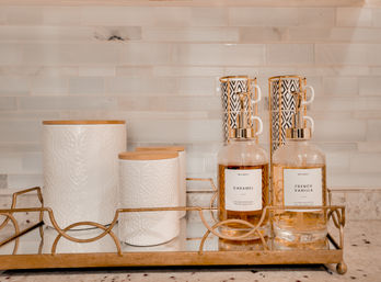 Kitchen countertop vignette: mirrored gold-trim tray with white textured ceramic canisters with bamboo lids, stacked black-and-white patterned mugs, and two gold-accented pump bottles labeled 'Caramel' and 'French Vanilla' against a subway tile backsplash.