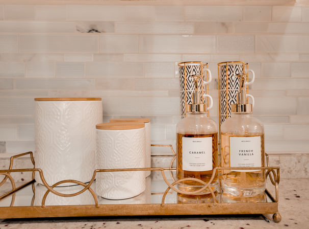 Kitchen countertop vignette: mirrored gold-trim tray with white textured ceramic canisters with bamboo lids, stacked black-and-white patterned mugs, and two gold-accented pump bottles labeled 'Caramel' and 'French Vanilla' against a subway tile backsplash.