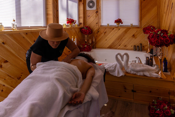 Cozy wood-paneled spa cabin: client receiving a back massage beside a jetted tub with towel swans, red rose bouquets, candles and soft lighting.
