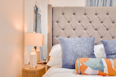 Cozy modern bedroom interior with beige tufted headboard, white bedding, blue patterned throw pillows and orange-teal bolster pillow, wooden round bedside table with textured ceramic lamp and wall mirror.