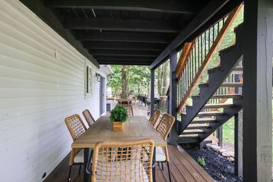 Cozy covered backyard porch with an outdoor dining table and wicker chairs under a dark-painted deck, wooden staircase to an upper deck, and a leafy wooded backyard view.