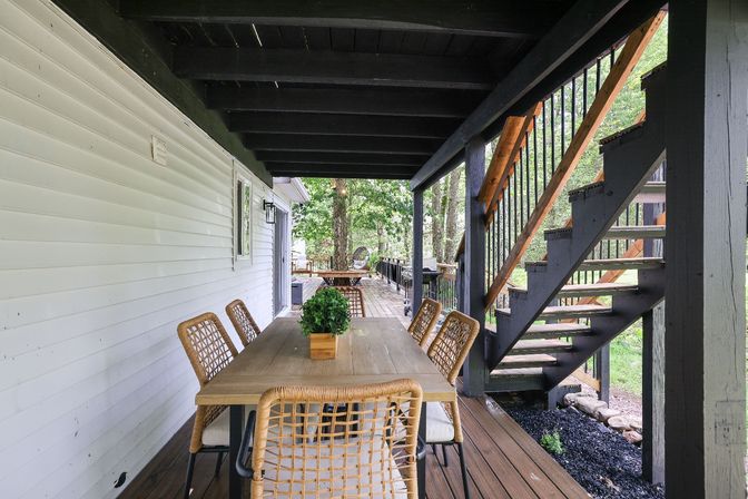 Cozy covered backyard porch with an outdoor dining table and wicker chairs under a dark-painted deck, wooden staircase to an upper deck, and a leafy wooded backyard view.