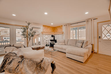 Cozy open-plan suburban living room with light beige sectional sofa, glass coffee table, wood-burning stove beneath a wood-paneled mantel, large windows and sliding doors, hardwood floors and neutral textured decor.
