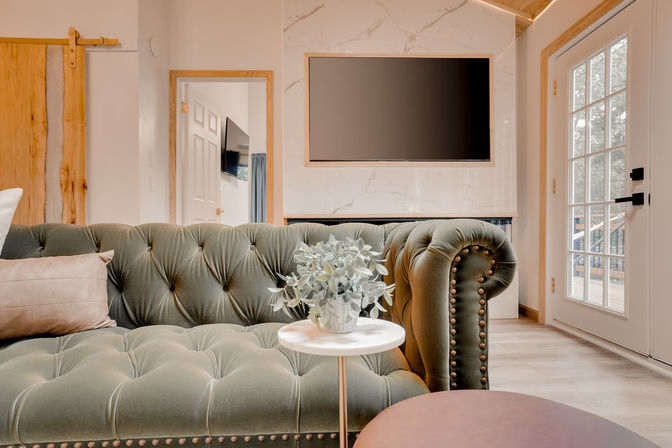 Cozy modern-rustic living room with a green tufted velvet Chesterfield sofa, round marble side table with potted plant, wall-mounted flat-screen on a marble accent wall, wood barn door and glass French door letting in natural light.