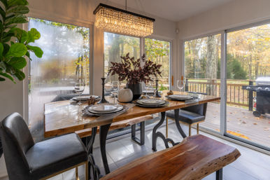 Modern rustic dining room with live-edge wood table set for four, crystal chandelier, leather chairs and bench, large windows and sliding glass doors overlooking an autumn wooded backyard and deck with barbecue