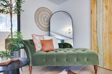 Sunlit modern living room corner with a green velvet chaise lounge, colorful throw pillows, arched floor mirror, woven boho wall art, indoor plants, and wood accents.