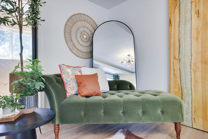 Sunlit modern living room corner with a green velvet chaise lounge, colorful throw pillows, arched floor mirror, woven boho wall art, indoor plants, and wood accents.
