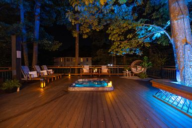 Nighttime backyard wooden deck with a glowing sunken hot tub, cushioned lounge chairs, a round rattan hanging chair, candles and lanterns, and blue uplighting on surrounding trees for a cozy outdoor retreat.