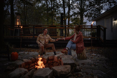 Couple enjoying a cozy autumn evening around a stone backyard firepit on a wooded deck patio, reaching hands across the fire as tiki torches glow and a pumpkin sits nearby at dusk.