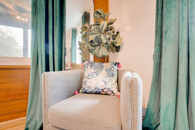 Cozy living room reading nook with a beige upholstered armchair, tropical-pattern throw pillow with pink tassels, green velvet curtains, fiddle-leaf fig plant and wooden accent panel.