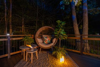 Cozy candlelit backyard deck at night with a round rattan lounge chair and tropical-print pillow, nested side tables with a small tabletop flame, potted ferns and a fiddle‑leaf fig, wood railing and dark trees beyond.