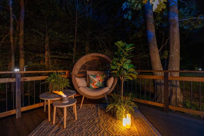 Cozy candlelit backyard deck at night with a round rattan lounge chair and tropical-print pillow, nested side tables with a small tabletop flame, potted ferns and a fiddle‑leaf fig, wood railing and dark trees beyond.