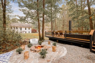 Cozy backyard stone fire‑pit patio with a glowing rock fire, log‑stump seats, potted ferns and a cushioned bench, next to a raised wooden deck surrounded by tall trees and a white two‑story house in a wooded suburban yard.