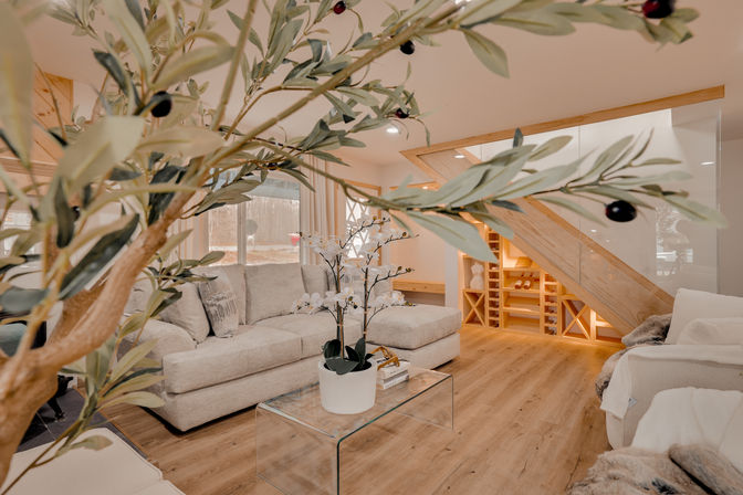 Airy modern cozy living room framed by leafy branches, featuring a neutral beige sectional, glass coffee table with potted orchids, light wood floors and built-in under-stair shelving and wine storage, softly lit by natural window light.