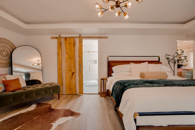 Modern rustic bedroom with a king-size bed dressed in white linens and green throw, sliding live-edge wooden barn door to an ensuite, tufted olive chaise with colorful pillows, large arched mirror, cowhide rug and brass multi-bulb chandelier.