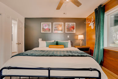 Cozy modern bedroom with a large metal-framed bed, dark gray accent wall and wood-paneled side, green velvet throw and pillows with a mustard bolster, three framed floral prints above the headboard, antler-style wall decor, bedside lamp with antler base, ceiling fan and a large window with green curtains.