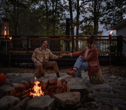 Couple enjoying a cozy autumn evening around a stone backyard firepit on a wooded deck patio, reaching hands across the fire as tiki torches glow and a pumpkin sits nearby at dusk.