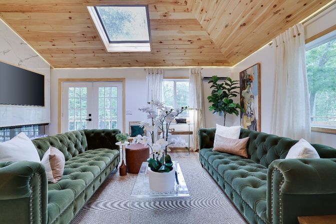 Bright modern living room with vaulted pine wood ceiling and skylight, twin green velvet tufted sofas facing a clear acrylic coffee table with white orchids, French doors, large windows, neutral rug and leafy plants