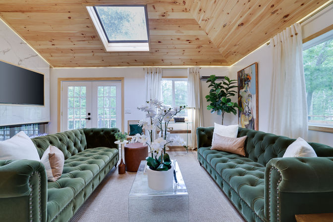 Bright cozy living room with twin green velvet tufted sofas, glass coffee table with white orchid centerpiece, wood-paneled vaulted ceiling with skylight, and French doors letting in natural light.