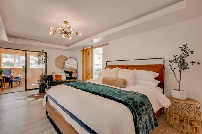 Bright modern boho master bedroom with cozy king bed in white linens and emerald green throw, leather headboard, gold globe chandelier, sliding glass doors to a sunroom, rattan side table and indoor plants