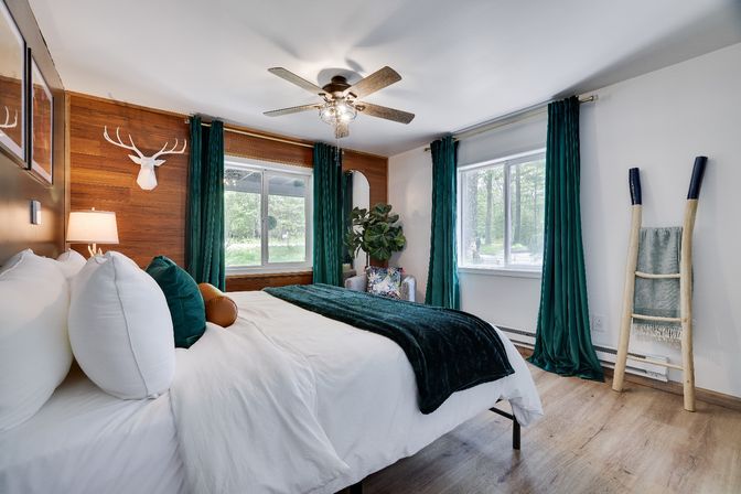 Cozy modern bedroom with wood accent wall, large windows looking out to trees, white bed with dark green velvet throw and pillows, teal curtains, ceiling fan, faux deer head wall decor, potted plant and ladder towel rack.