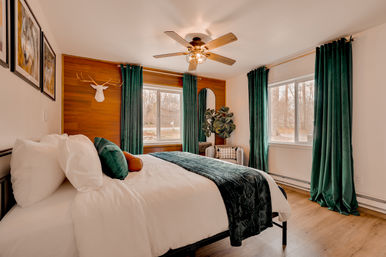 Cozy modern bedroom with white bedding, emerald green velvet curtains and throw, wood accent wall with decorative deer head, ceiling fan, corner plant and windows overlooking a wooded yard.