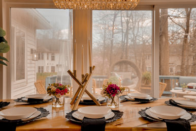 Cozy suburban dining table set for six by sliding glass doors overlooking a wooded backyard deck, with white plates on black placemats, black napkins, small floral arrangements and a modern gold candleholder centerpiece under a crystal chandelier.