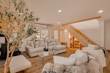 Cozy modern basement living room with neutral beige sectional, glass coffee table and potted orchid, indoor olive tree, wood-burning stove, warm wood floors and a wooden staircase with glass railing and built-in wine storage.