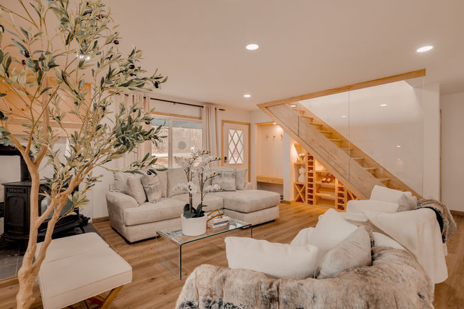 Cozy modern basement living room with neutral beige sectional, glass coffee table and potted orchid, indoor olive tree, wood-burning stove, warm wood floors and a wooden staircase with glass railing and built-in wine storage.