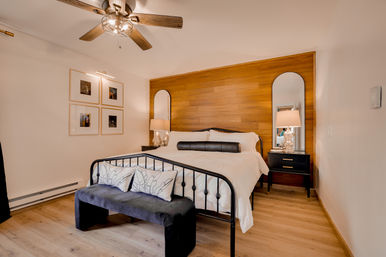 Cozy modern bedroom with warm wood accent wall, black metal-framed bed dressed in white linens, arched mirrors, matching black nightstands with lamps, velvet bench at the foot, ceiling fan, and light hardwood floor.